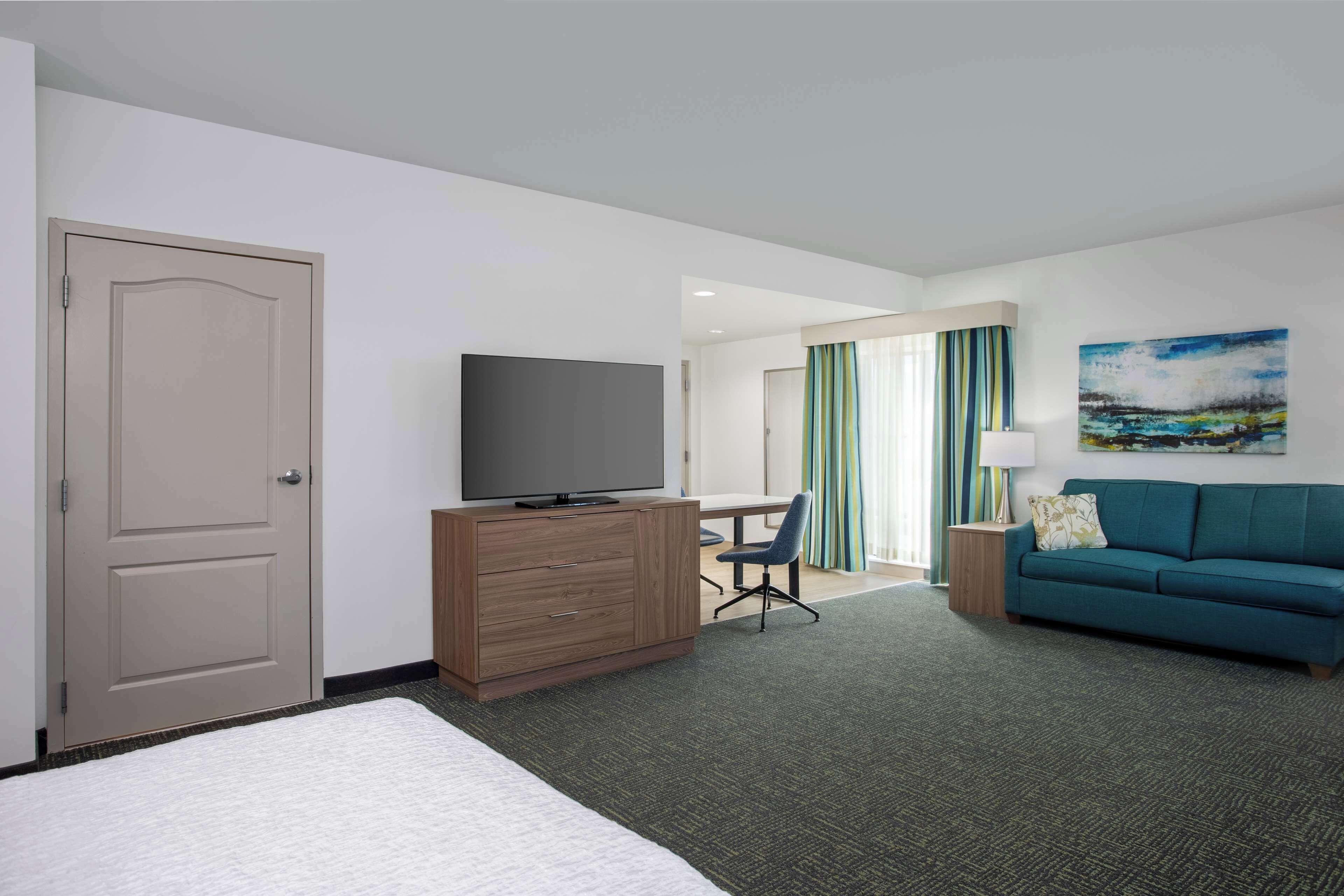 A hotel room with a bed, a TV on a wooden dresser, a desk with a chair, a teal sofa, and a painting on the wall.