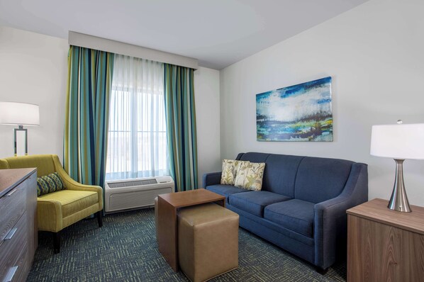 Una habitación de hotel con un sofá azul, un sillón amarillo, una mesita de madera, una lámpara y un cuadro en la pared.