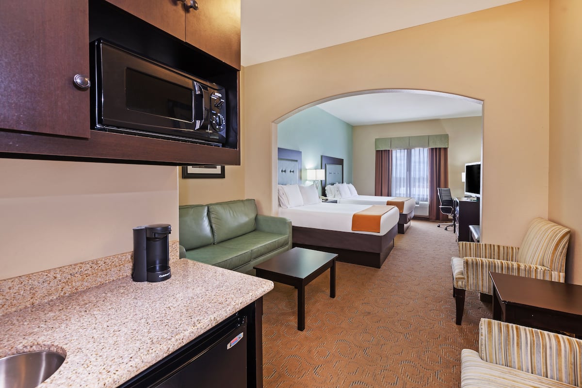 A hotel room with a kitchenette, a green sofa, a bed with two pillows, a desk, and a striped armchair.