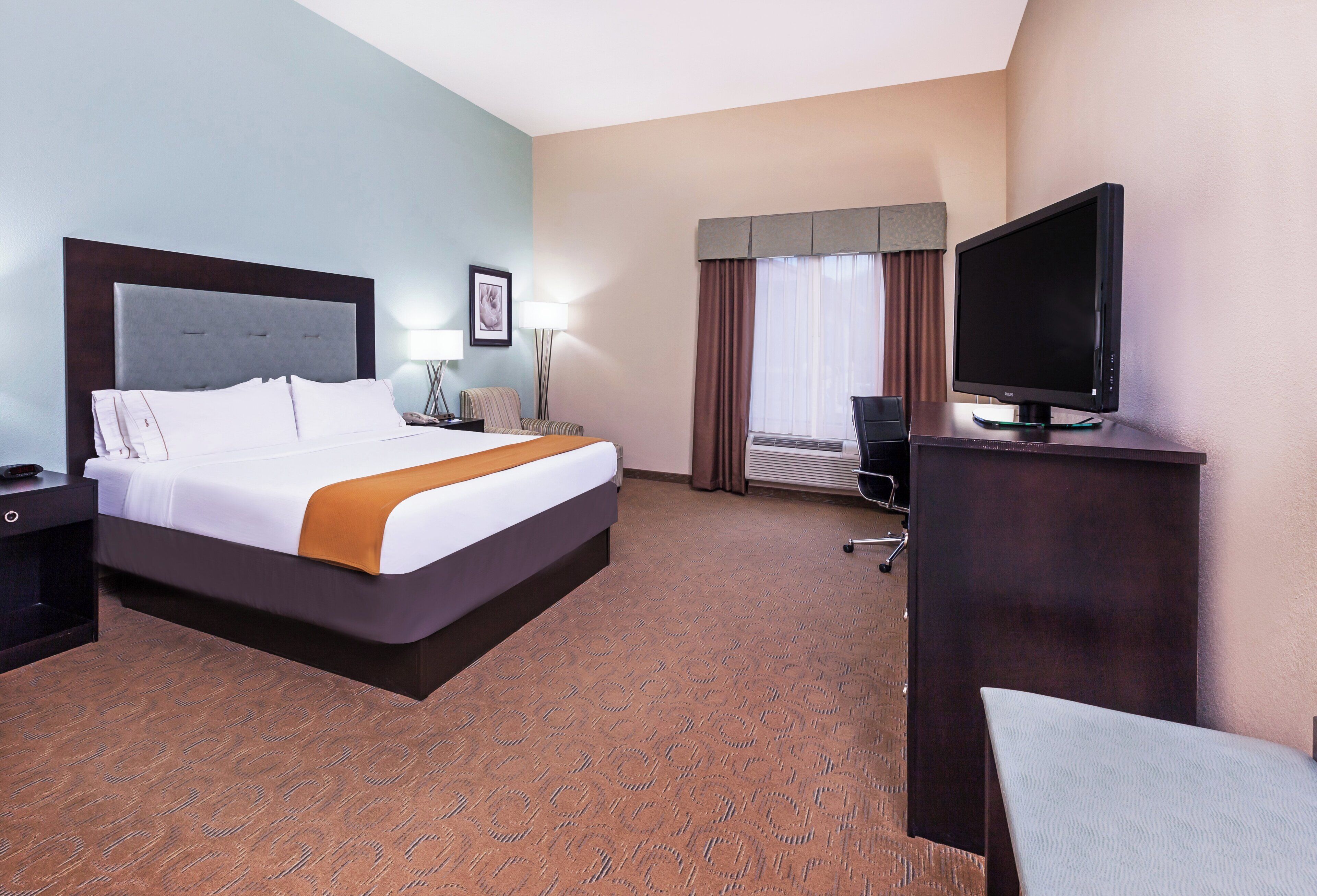 A hotel room with a bed, a desk with a television, a chair, a lamp, and a picture on the wall.