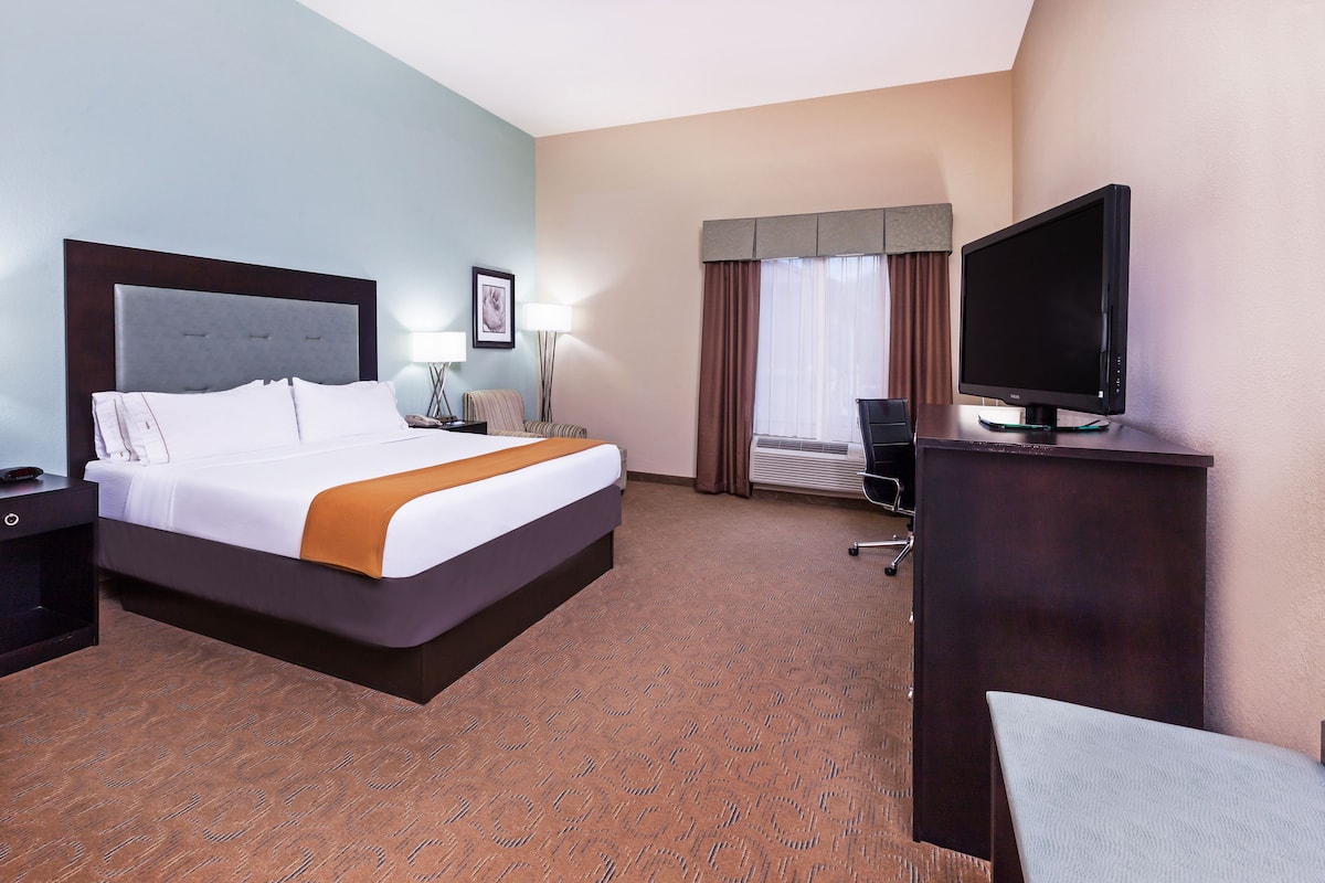 A hotel room with a large bed, a desk with a flat-screen TV, a chair, a lamp, and a window with curtains.