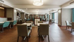 Un amplio comedor de hotel con numerosas mesas y sillas, una zona de bar central y grandes ventanales que dejan entrar luz natural.