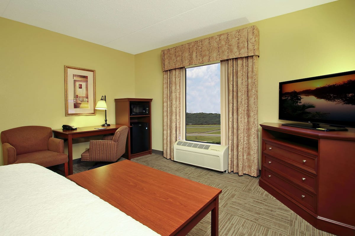 A hotel room with a bed, a desk with a lamp, a television, a chair, a dresser, and a large window with curtains.