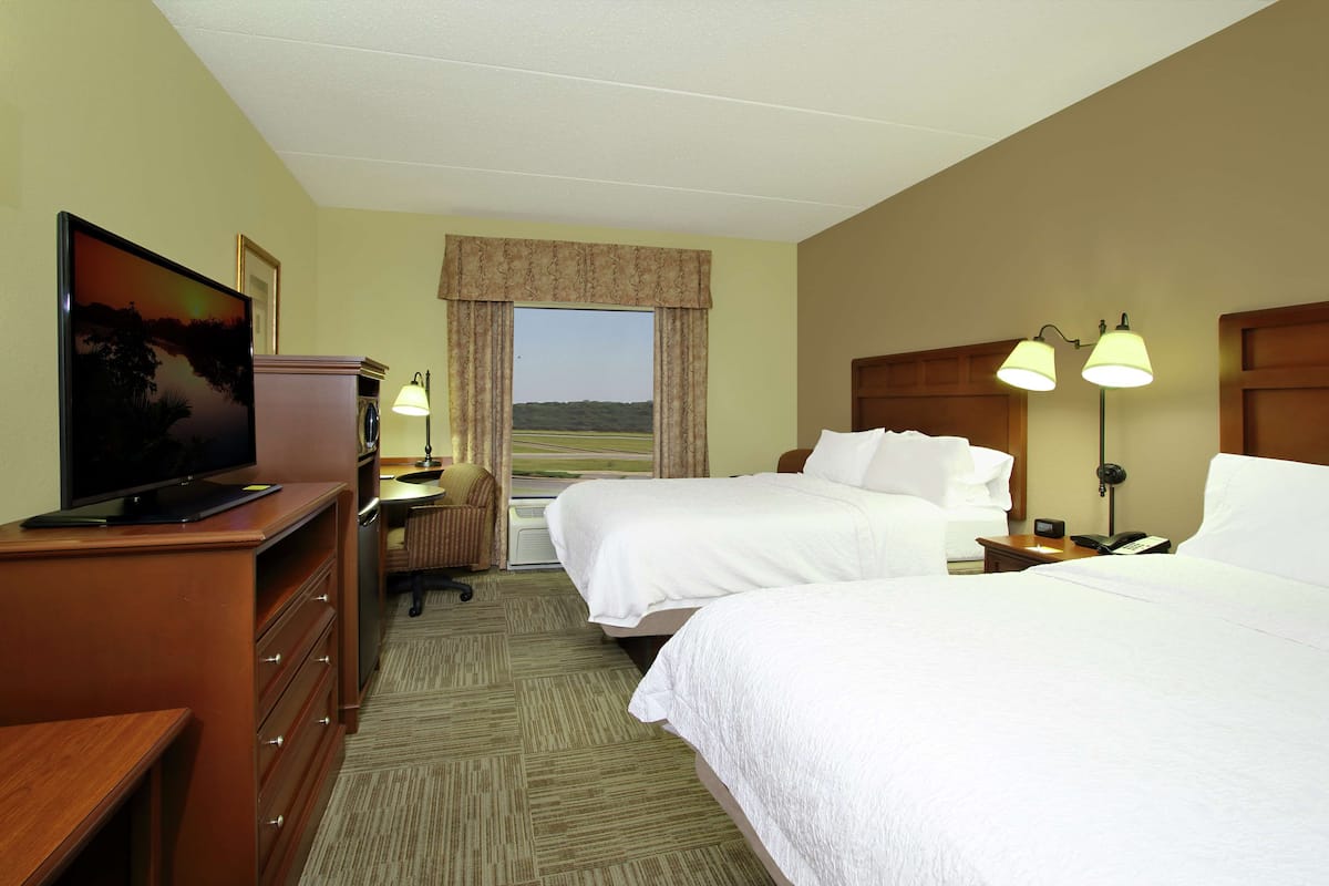 A hotel room with two beds, a television, a desk with a lamp, and a view of the outdoors.