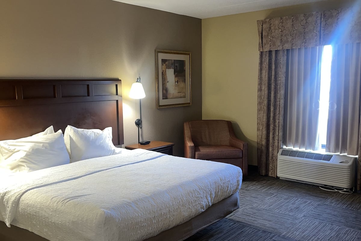 A hotel room with a large bed, a chair, a painting on the wall, and a window with curtains.
