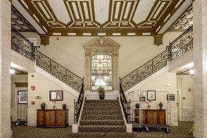 Una imponente escalera con barandillas ornamentales y un arco decorativo central, que conduce a una zona de descanso con una planta en maceta y obras de arte enmarcadas.