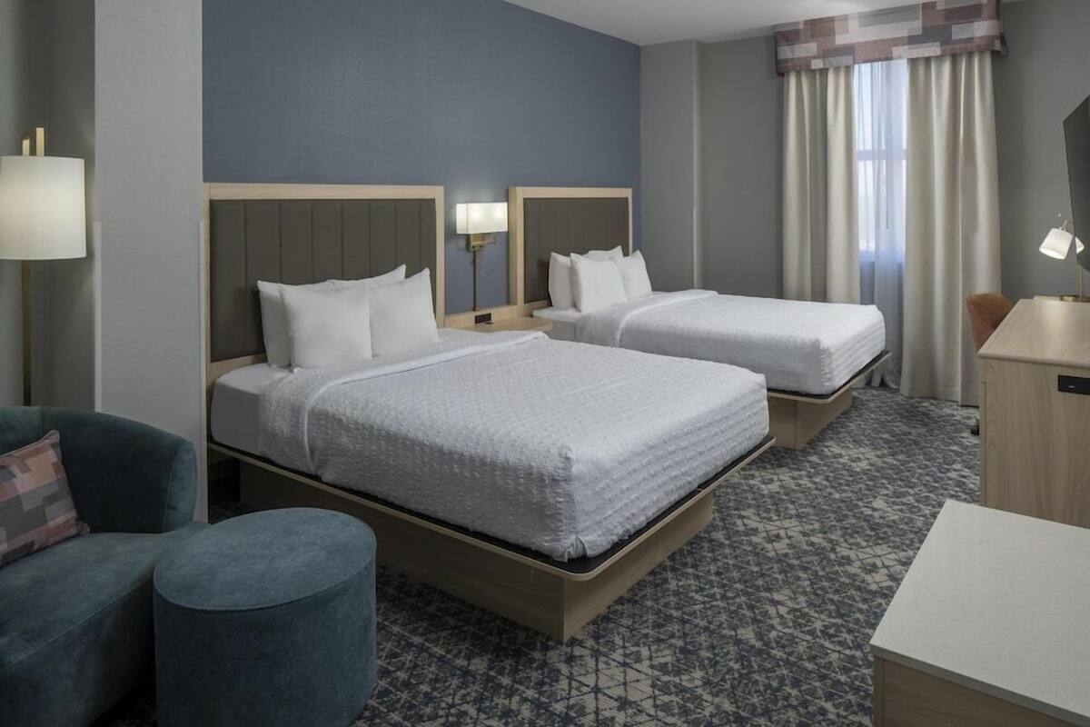 A hotel room with two beds, a desk, a chair, and a lamp.