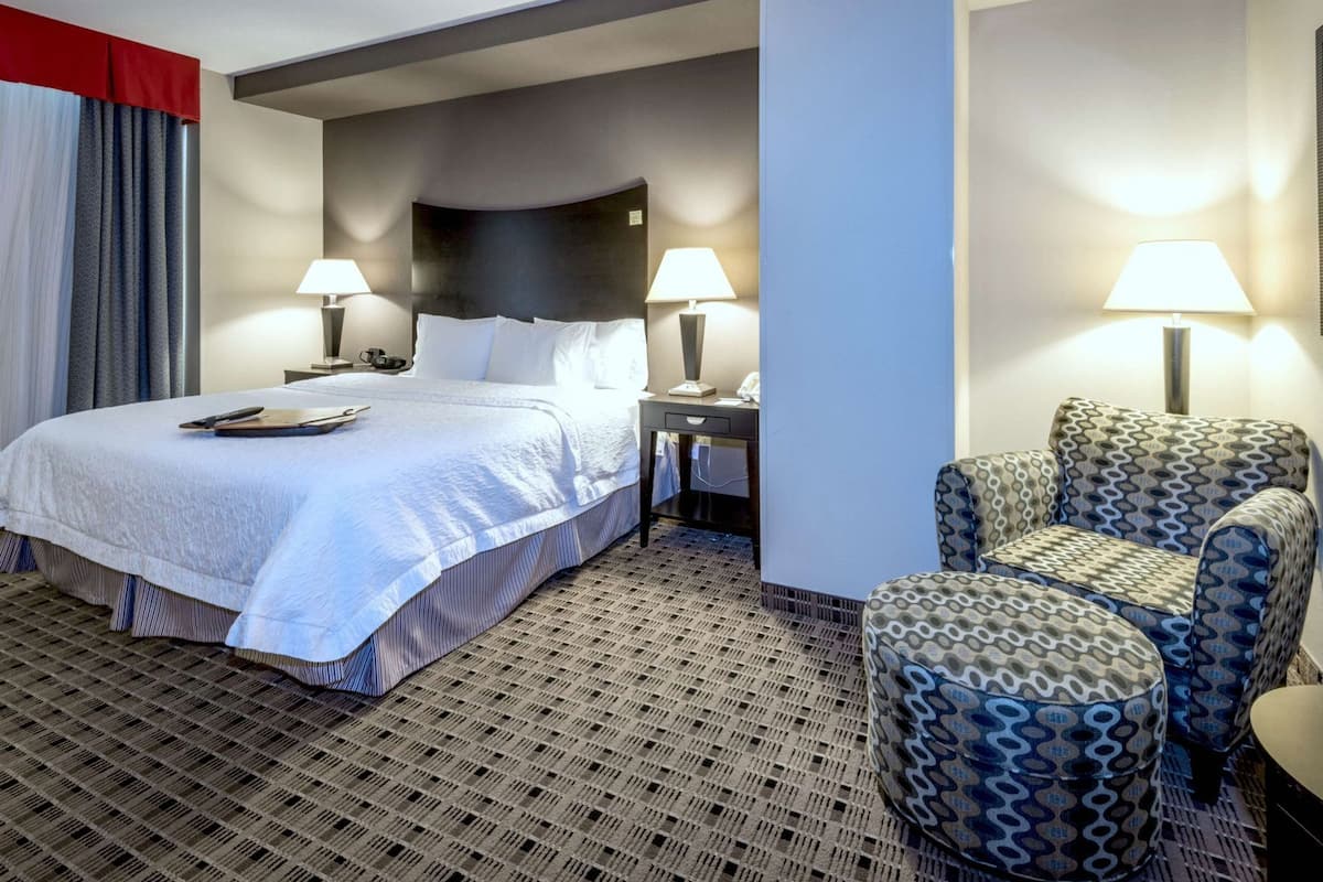 A hotel room with a bed, two bedside lamps, a desk with a telephone, a patterned armchair, and a patterned carpet.