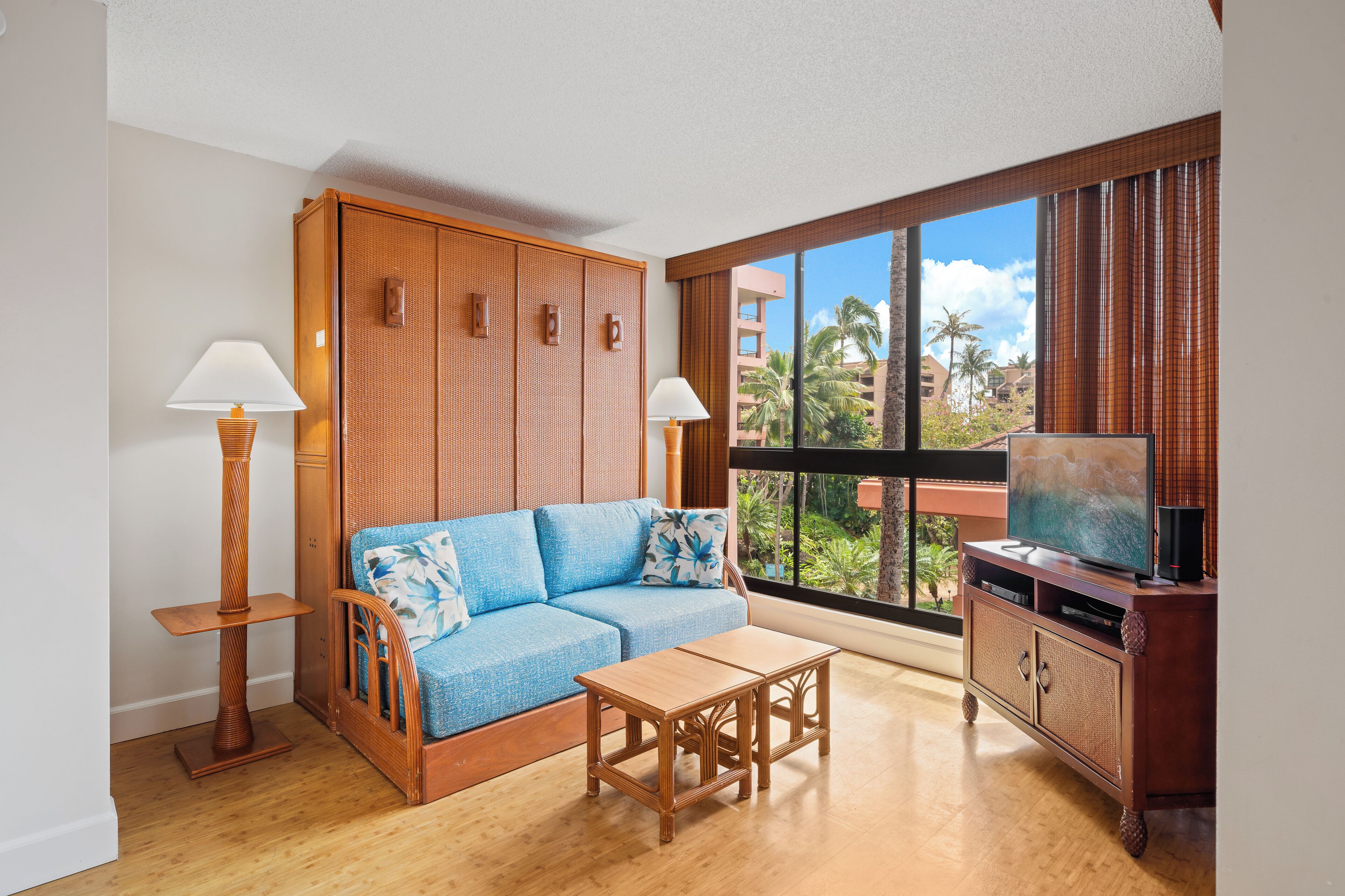 A living room with a blue sofa, a wooden coffee table, a TV on a wooden cabinet, and a view of palm trees through large windows.