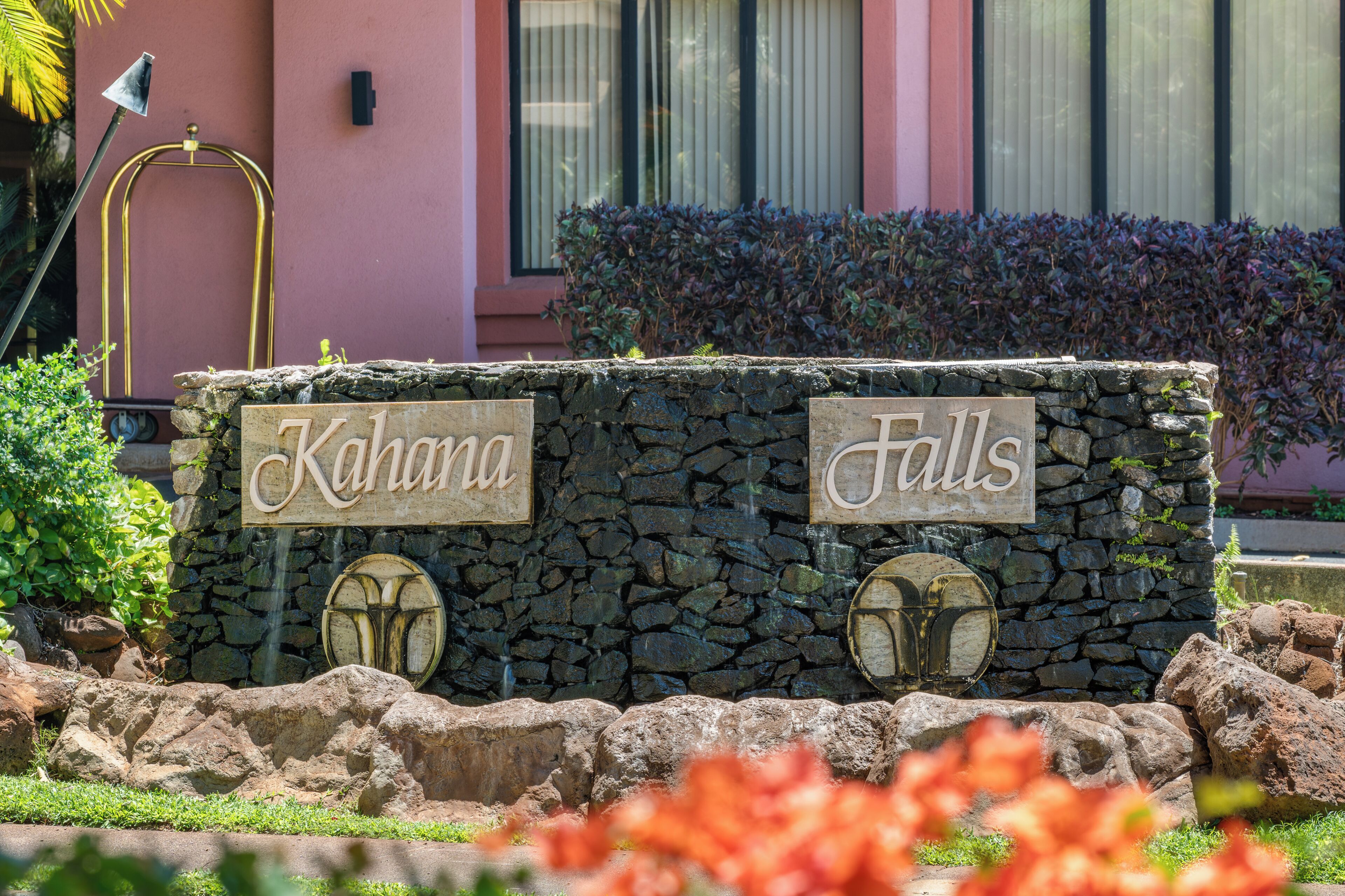 A stone wall with "Kahuna Falls" signs, surrounded by rocks and plants.