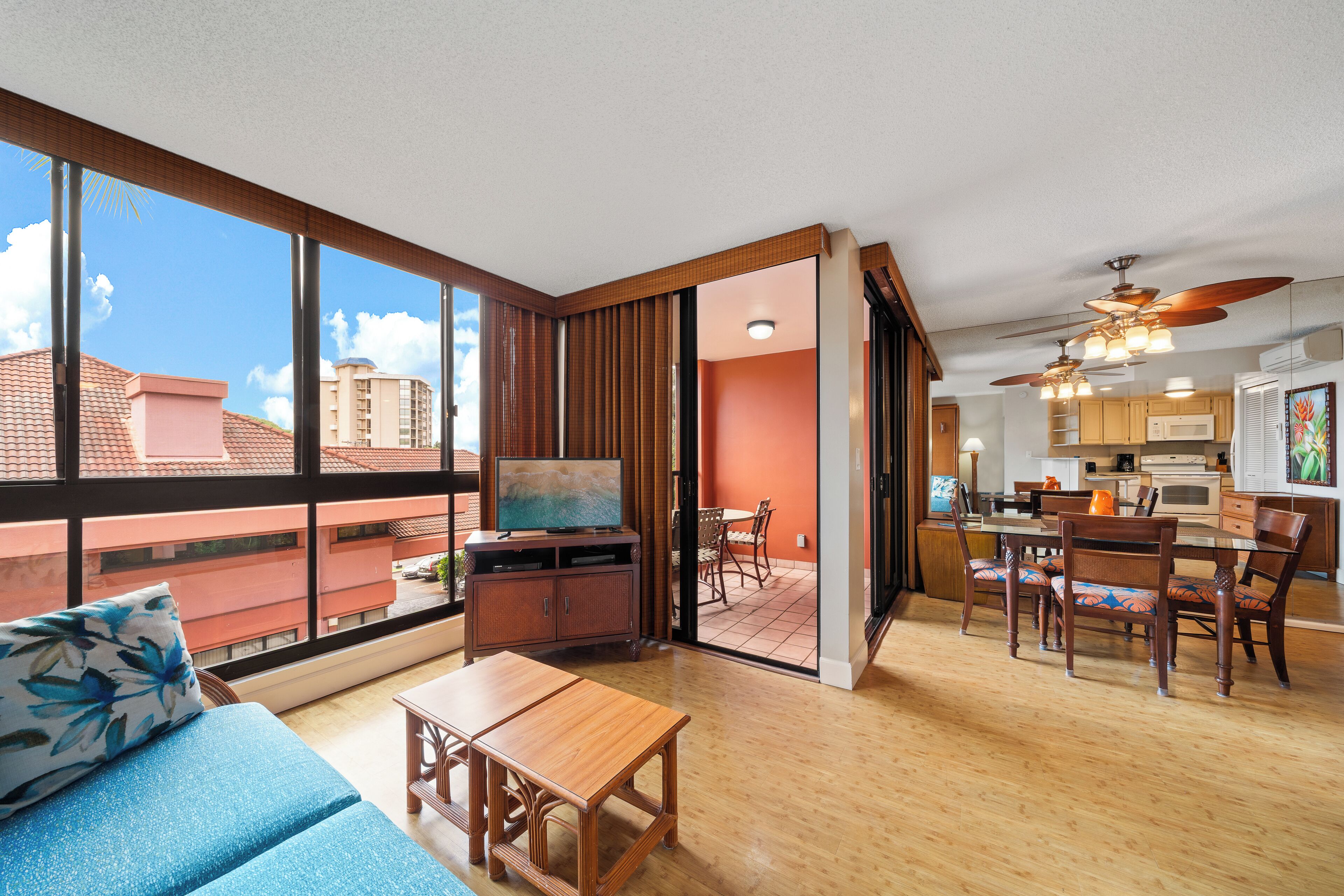 A spacious living area with a large window, a dining table, and a kitchen in the background.