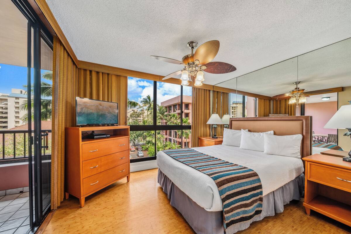 A hotel room with a large bed, wooden furniture, a television, and a balcony with a view of palm trees and buildings.
