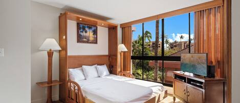 Habitación de hotel con una cama grande, un televisor sobre un mueble de madera y vistas a palmeras y edificios.