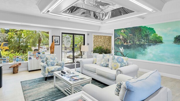 A spacious living room with a large window, a white sofa, a coffee table, and a painting of a tropical scene. - Anchorage Resort & Yacht Club (Key Largo)