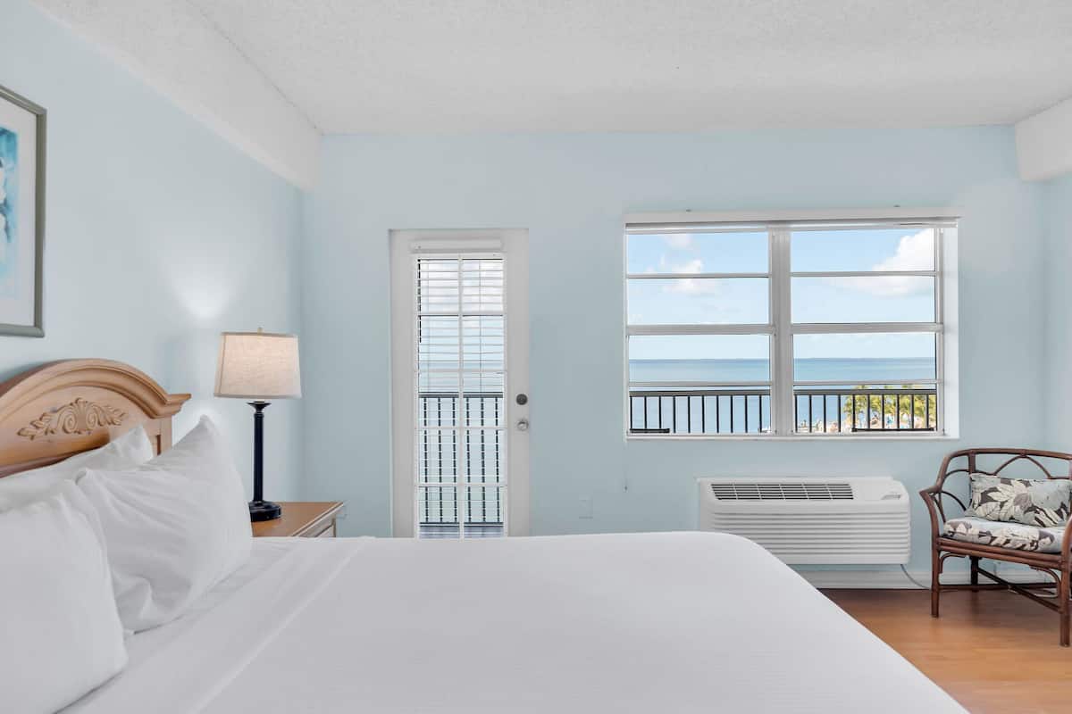 A bedroom with a bed, a nightstand, a lamp, a window with a view of the sea, and a chair.