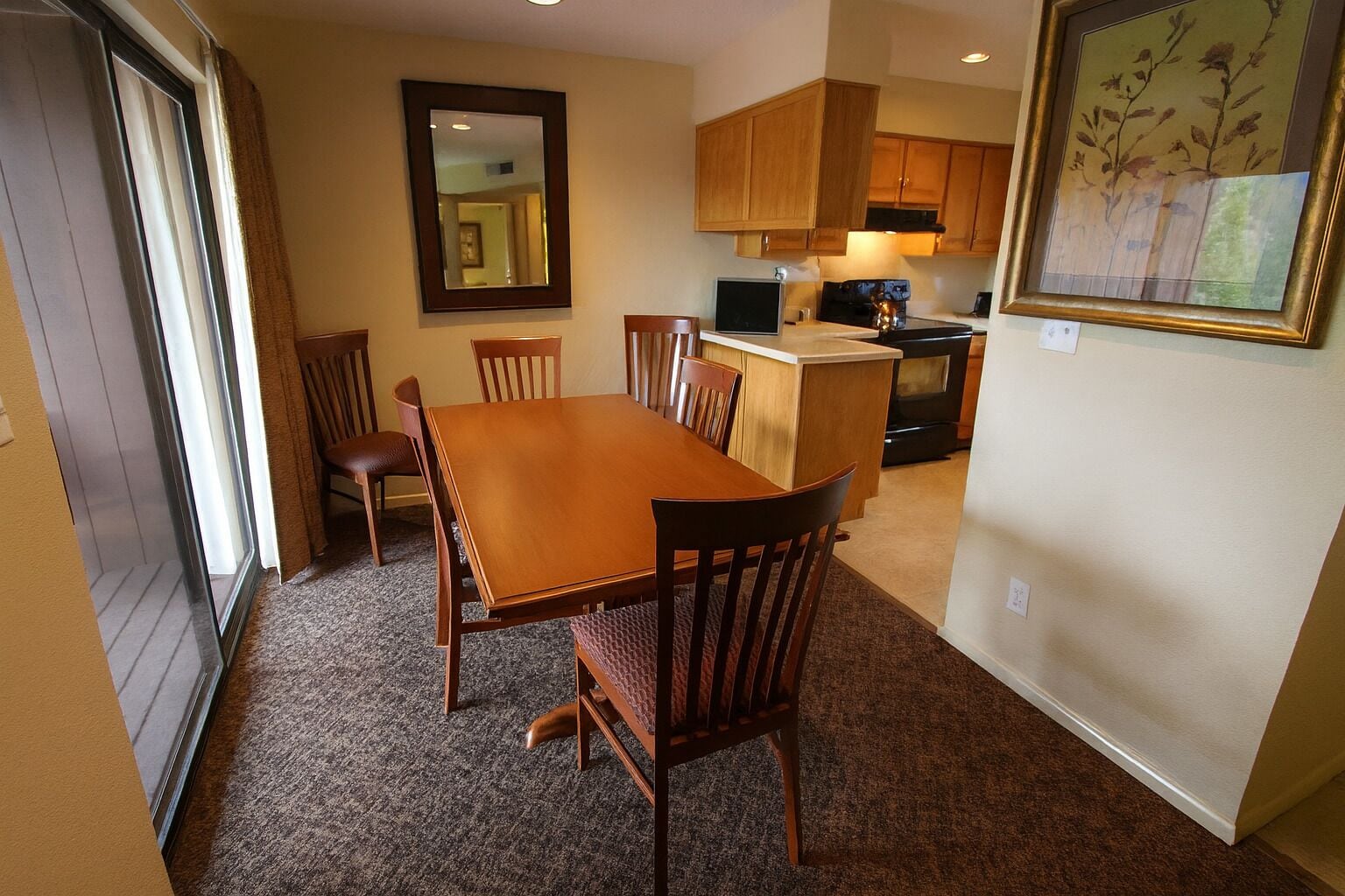 A dining area with a wooden table and chairs, a kitchen with wooden cabinets, a large mirror, and a framed artwork on the wall.