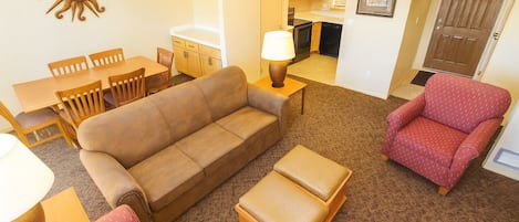 Ein Wohnzimmer mit einer Couch, Sesseln, einem Hocker, einem Esstisch mit Stühlen, einer Lampe und einer Küchenzeile im Hintergrund.