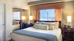 A hotel room with a large bed, a nightstand with a lamp, a mirror, and a view of the ocean.