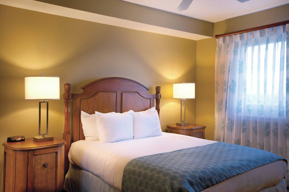 Ein Hotelzimmer mit einem hölzernen Kopfende, zwei Nachttischlampen, einem Bett mit weißer Bettwäsche und einem Fenster mit durchsichtigen Vorhängen.