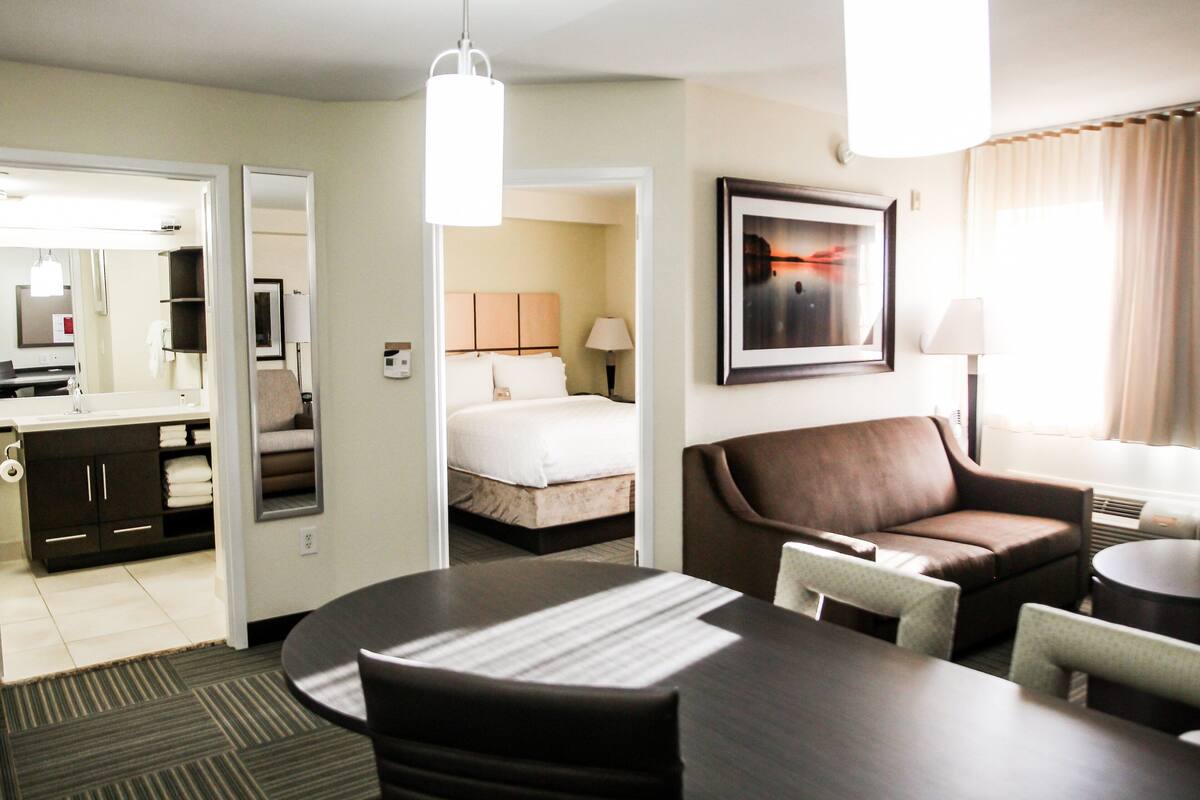 A hotel room with a bed, a brown leather sofa, a dining table with chairs, a small round table, a mirror, a framed picture on the wall, and a window with curtains.