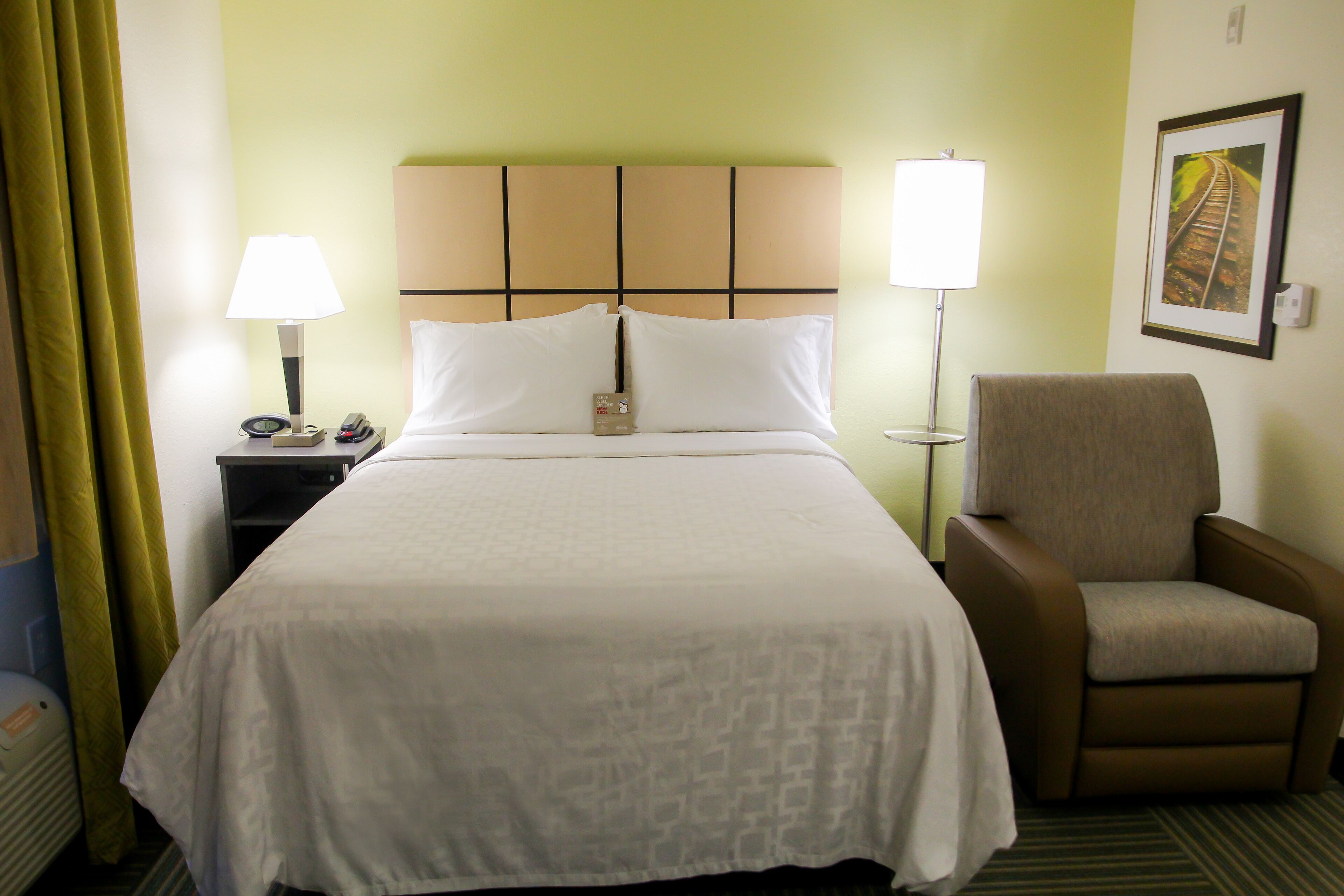 A hotel room with a bed, two bedside lamps, a chair, a nightstand, and a framed picture on the wall.