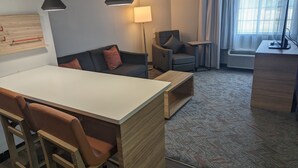 A modern hotel room with a white desk, two chairs, a sofa with cushions, a standing lamp, a side table, a TV, and a large window with curtains. - Candlewood Suites Wilson by IHG (Wilson)
