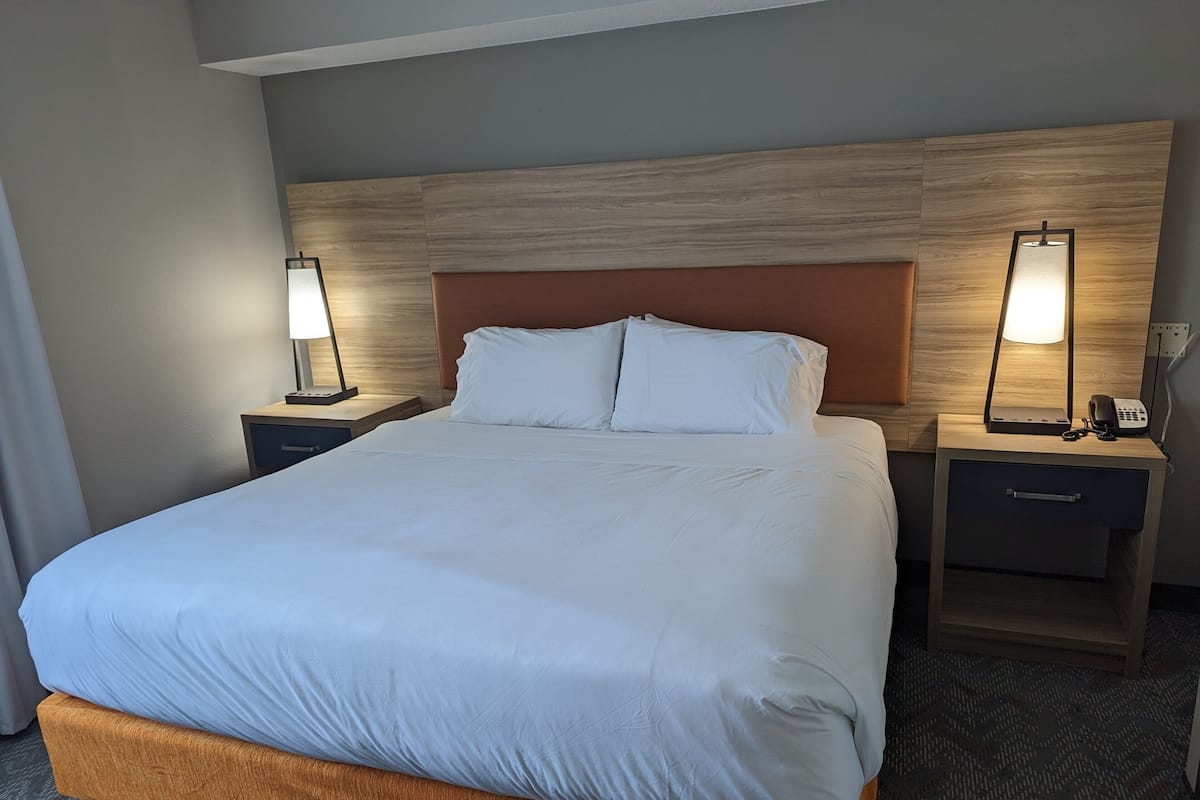 Hotellihuone, jossa on suuri sänky, kaksi yöpöytää valaisimineen, puinen pääty ja matolla peitetty lattia.