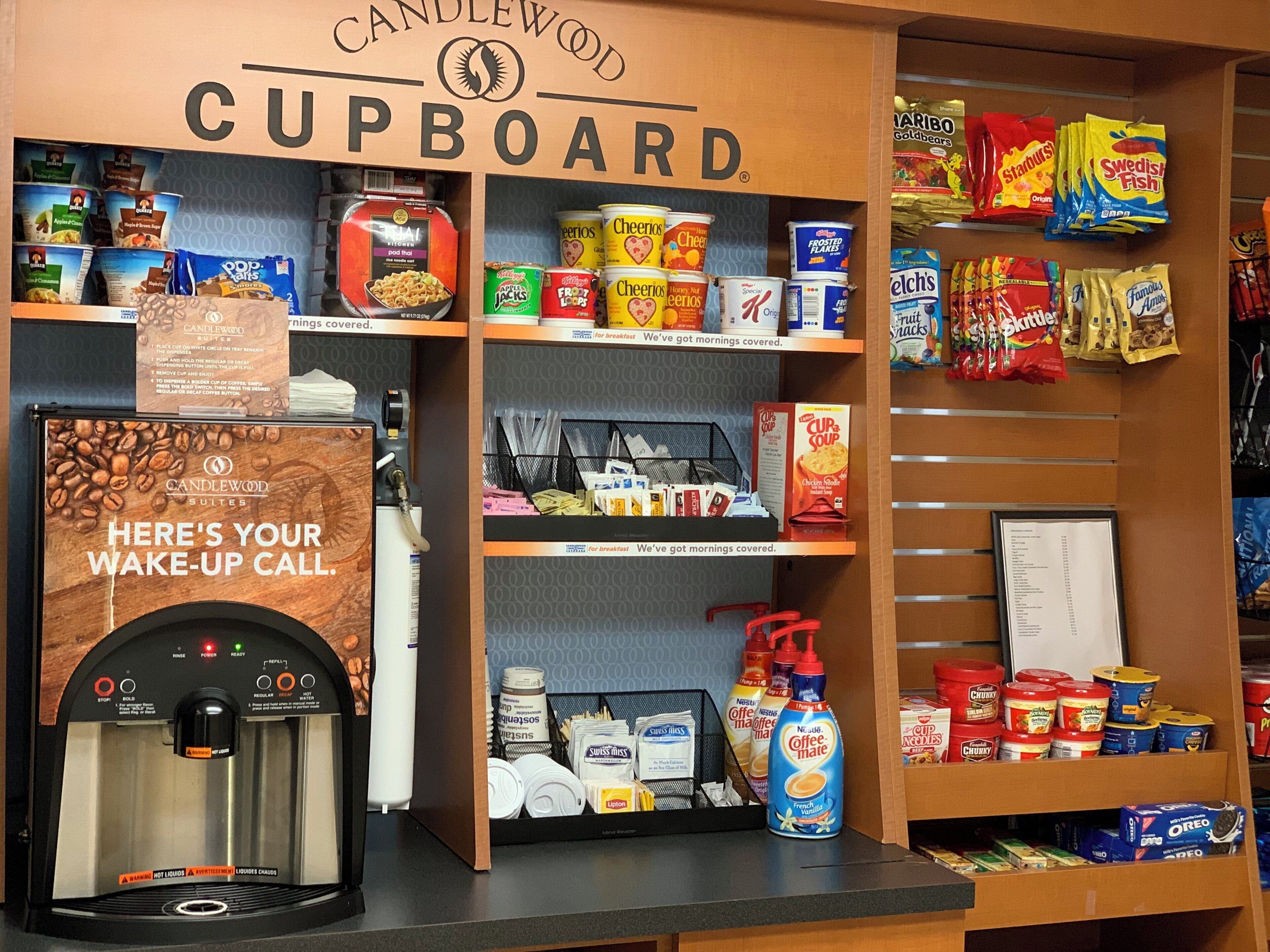 Un coin cuisine équipé d’une machine à café, d’étagères remplies de produits pour le petit-déjeuner et d’un panneau indiquant la disponibilité d’un service de réveil.