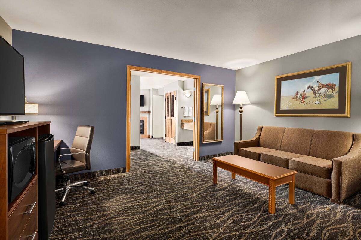 Ein Hotelzimmer mit einem großen Spiegel, einem braunen Sofa, einem Holztisch, einem Fernseher und einem Schreibtisch mit einem Computer.