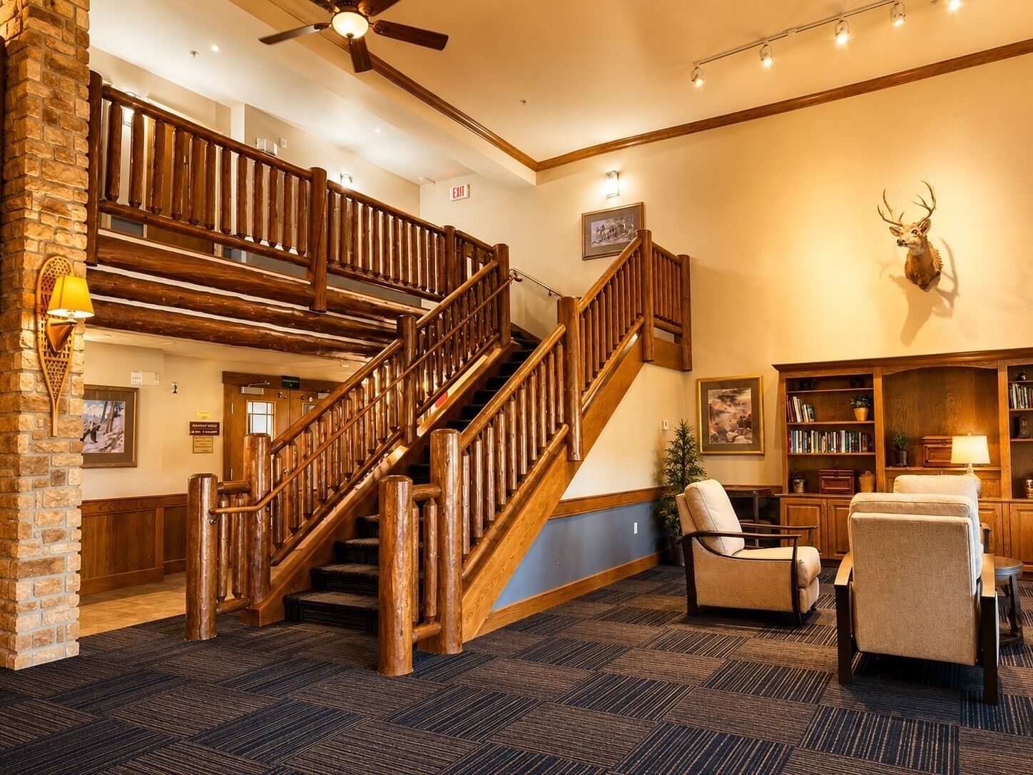 A lobby with a wooden staircase, a fireplace, a bookshelf, and a mounted deer head.