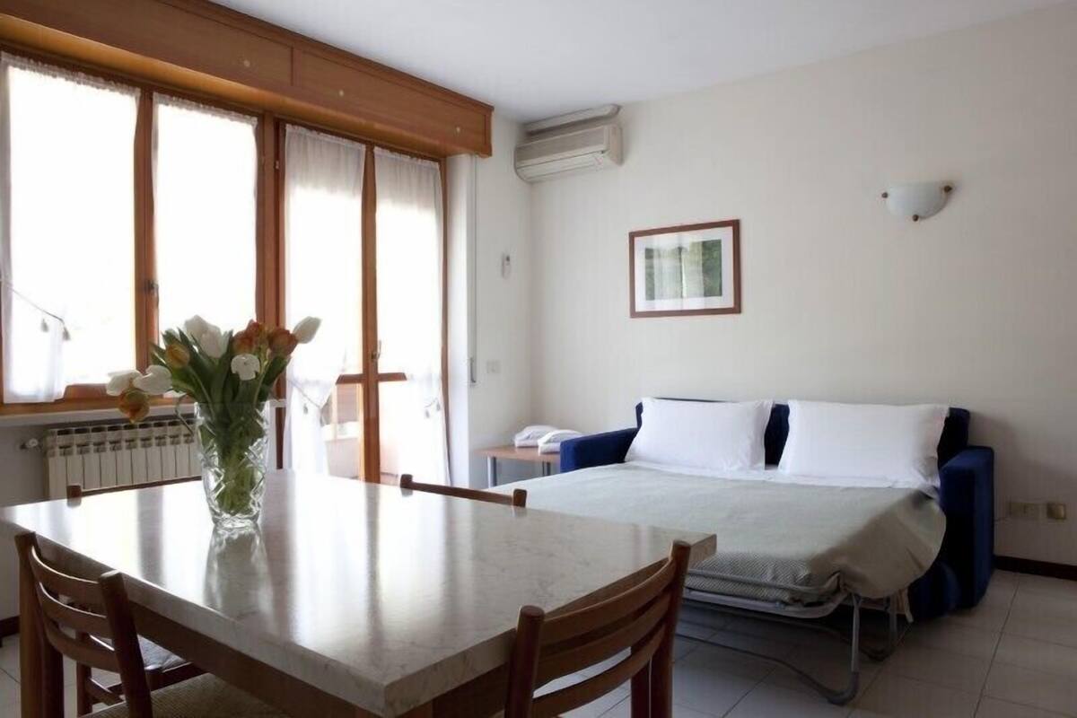 A hotel room with a bed, a table with chairs, a vase with flowers, a radiator, and a window with curtains.
