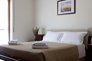 Uma cama bem arrumada com lençóis brancos e um espalha branco, um quadro na parede e um vaso com flores secas em uma mesa de cabeceira.