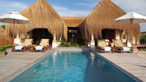 A pool area with two thatched-roof structures, lounge chairs, and umbrellas. - Azura Benguerra Island (Benguerra Island)