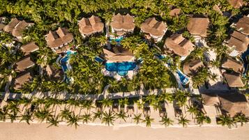Vista aérea de un resort con varias edificaciones de techo de paja, una piscina central y palmeras bordeando la playa.