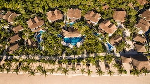 An aerial view of a resort with multiple thatched-roof structures, a central swimming pool, and palm trees lining the beachfront.