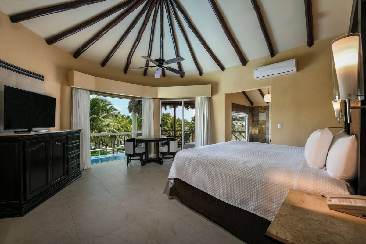 A bedroom with a large bed, a ceiling fan, a TV, a balcony with a table and chairs, and a view of palm trees.