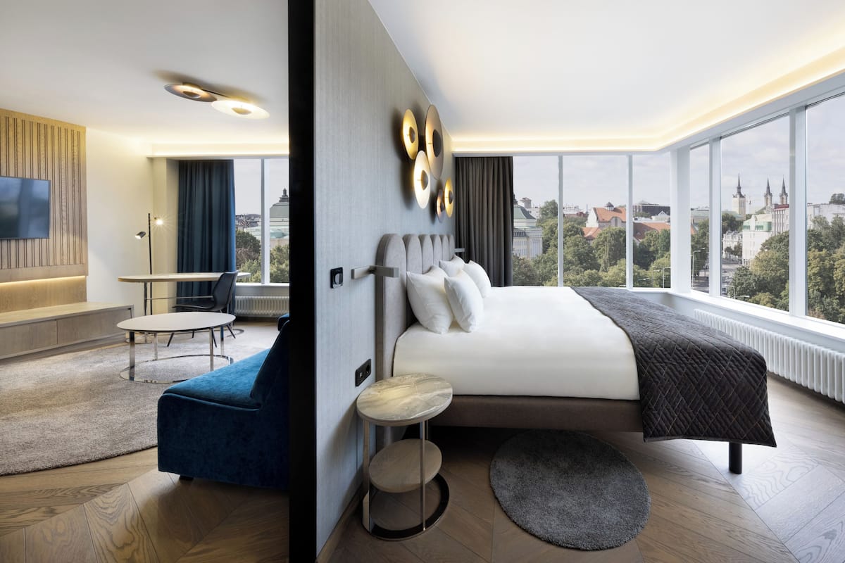 Une chambre d’hôtel moderne avec un grand lit, un coin salon comprenant un fauteuil bleu et une petite table ronde, et une vue sur la ville grâce à de grandes fenêtres.