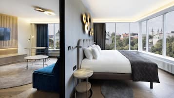 Habitación de hotel moderna con una cama grande, una zona de estar con una silla azul, una pequeña mesa redonda y vistas de la ciudad a través de amplios ventanales.