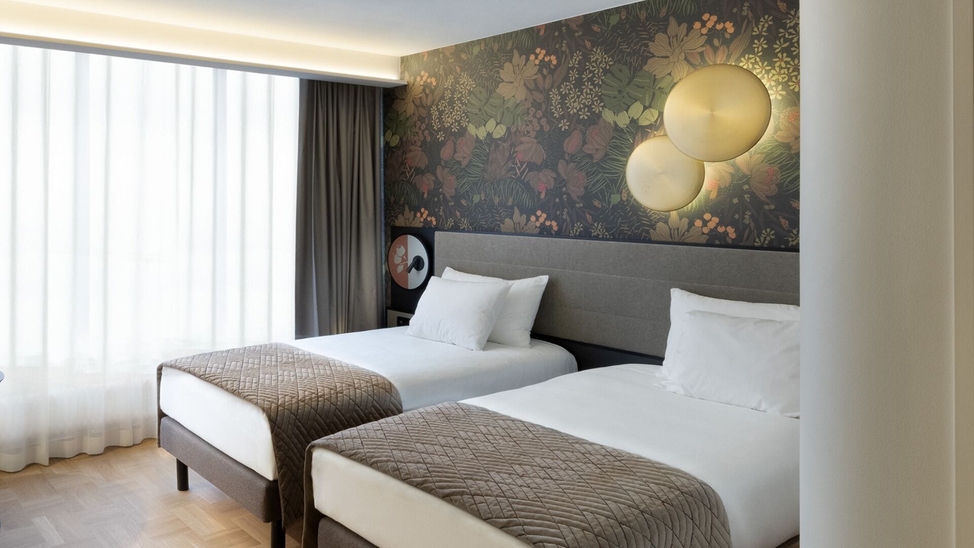 Habitación de hotel con dos camas, un ventanal con cortinas, un mural floral en la pared y un reloj de pared redondo.
