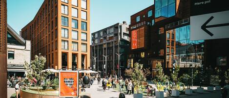 Una plaza urbana moderna con edificios residenciales y comerciales, un espacio central con asientos y una señal de indicaciones.
