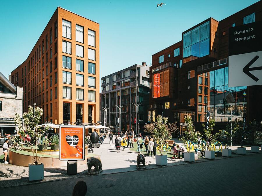 A modern urban square with a mix of commercial and residential buildings, a central plaza with seating, and a directional sign.