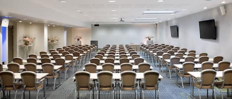 Un salón de conferencias con filas de mesas y sillas, un televisor de pantalla plana en la pared y luces en el techo.