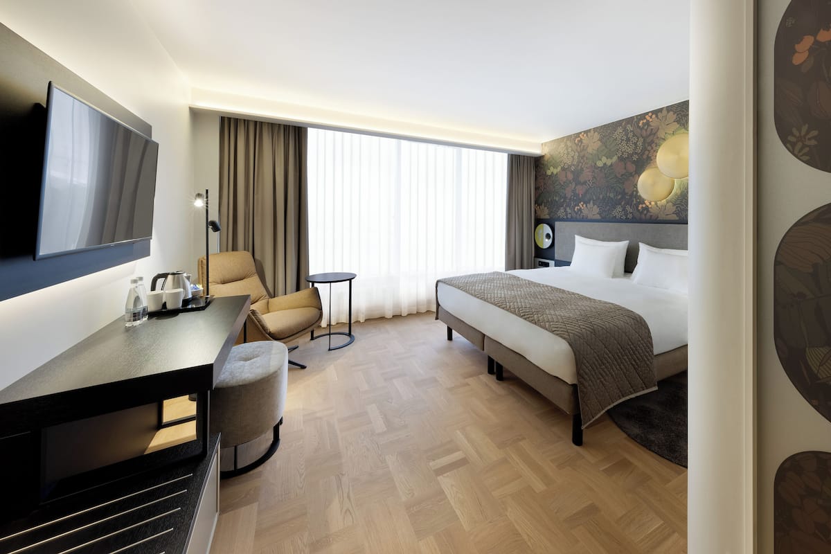 Une chambre d’hôtel avec un lit, un bureau, une chaise, une télévision et une fenêtre avec des rideaux.