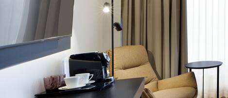 A hotel room with a dark wooden desk, a beige armchair, a small round table, and a floor lamp.