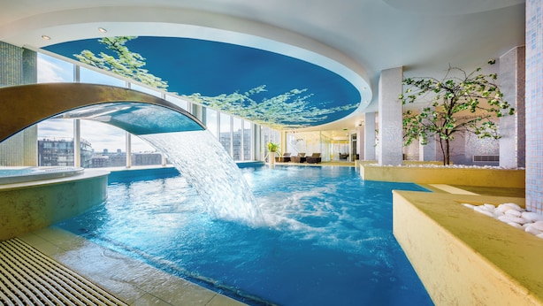 Una piscina interior con caída de agua, ventanas amplias y vistas a la ciudad.
