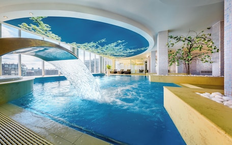 An indoor swimming pool with a waterfall feature, large windows, and a view of the cityscape.