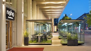 Un área de estar moderna al aire libre con mesas y sillas, rodeada de paredes de vidrio, y un letrero de restaurante a la vista.
