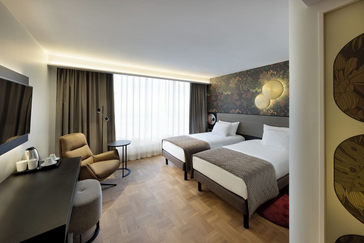 Une chambre d’hôtel avec deux lits, un bureau avec une chaise, une télévision et une grande fenêtre avec des rideaux.