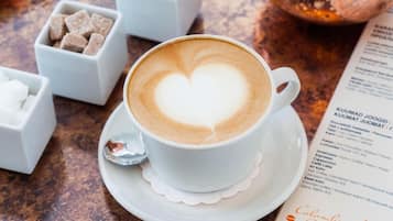 Una taza de café con dibujo de un corazón en la espuma, sobre un platito, acompañada de pequeños cuencos con azúcar y granos de café, y un menú sobre una mesa de madera.