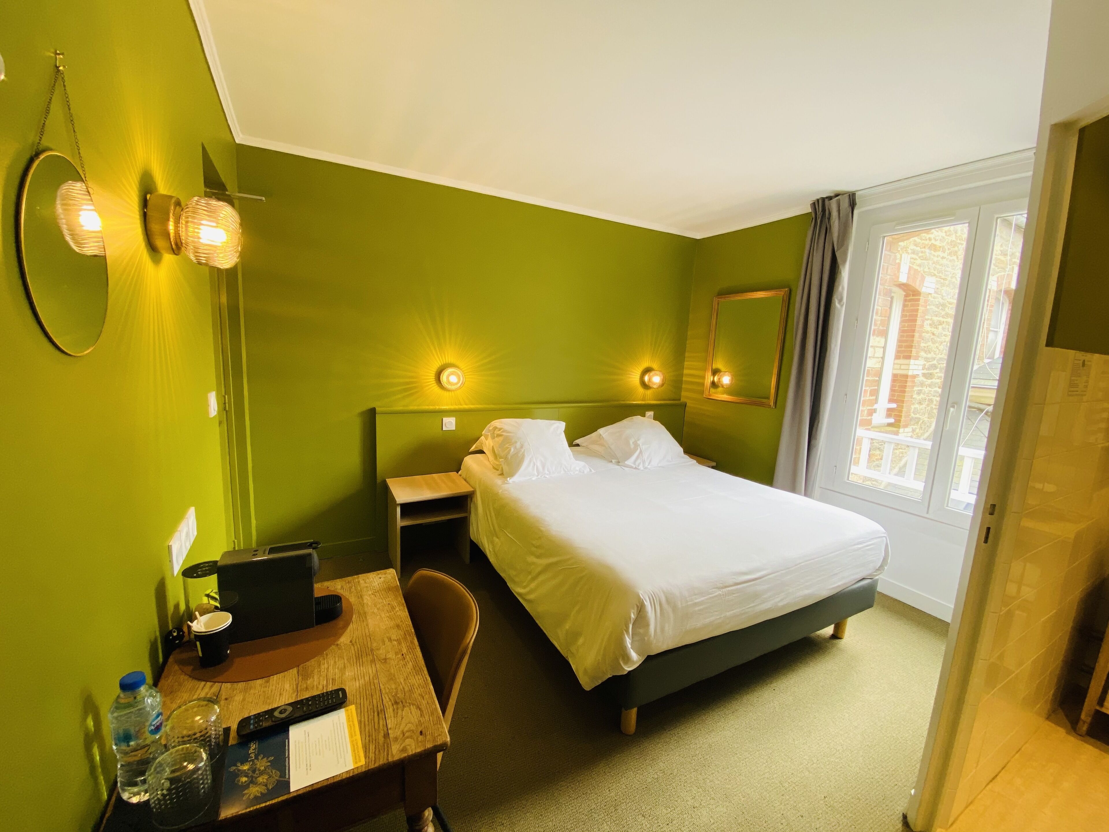 A hotel room with a bed, bedside tables, a desk with a laptop, a mirror, and a window with curtains.